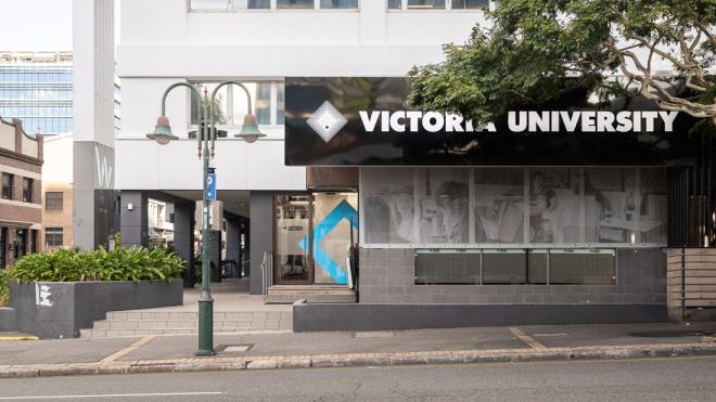 A photo of a street with the sign "Victoria University" on the building. A lamp with a parking sign attached to it is standing on the street.