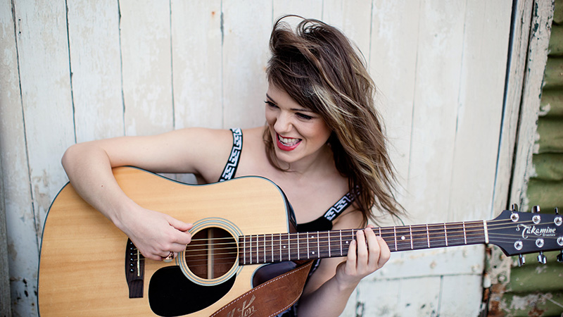 Young white woman plays acoustic guitar, smiling and singing, outdoors