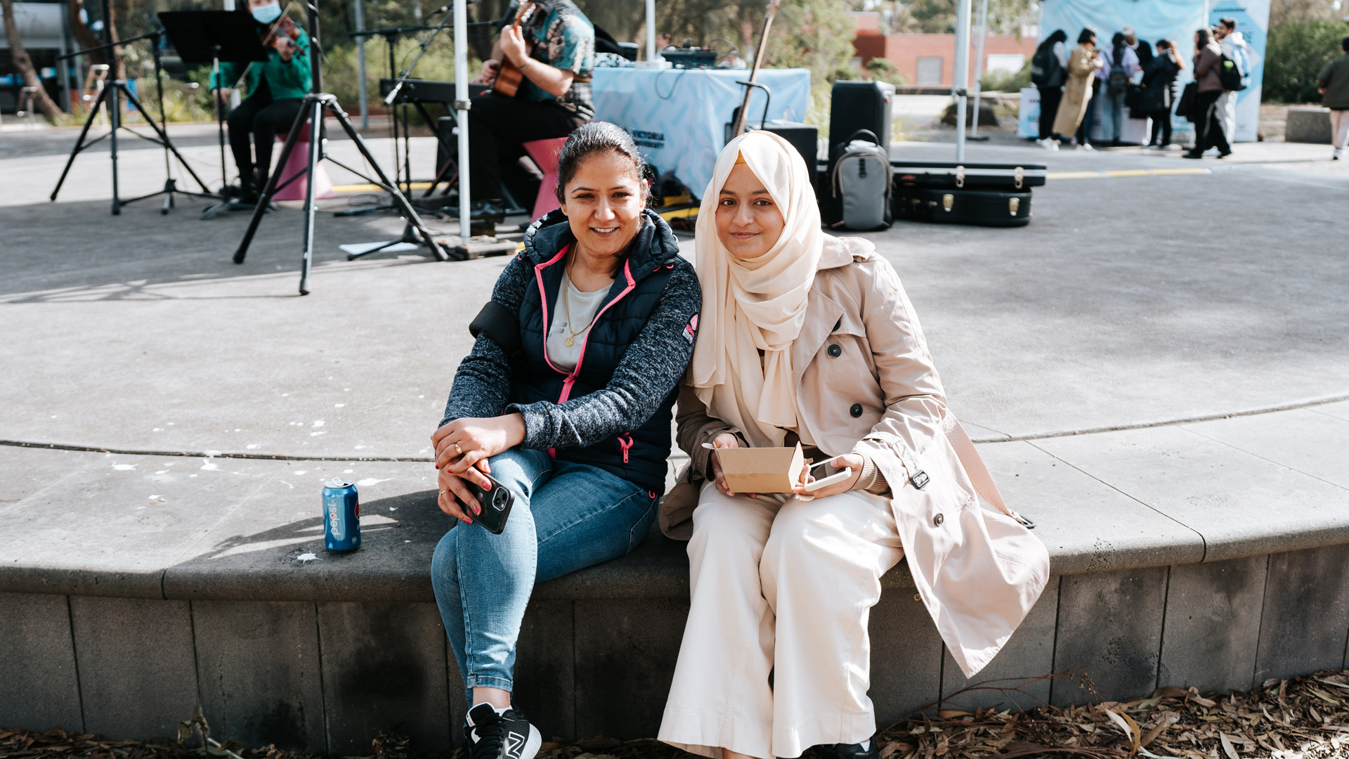 Two students sitting and smiling at camera. In the background are two musicians on chairs playing a guitar and violin.