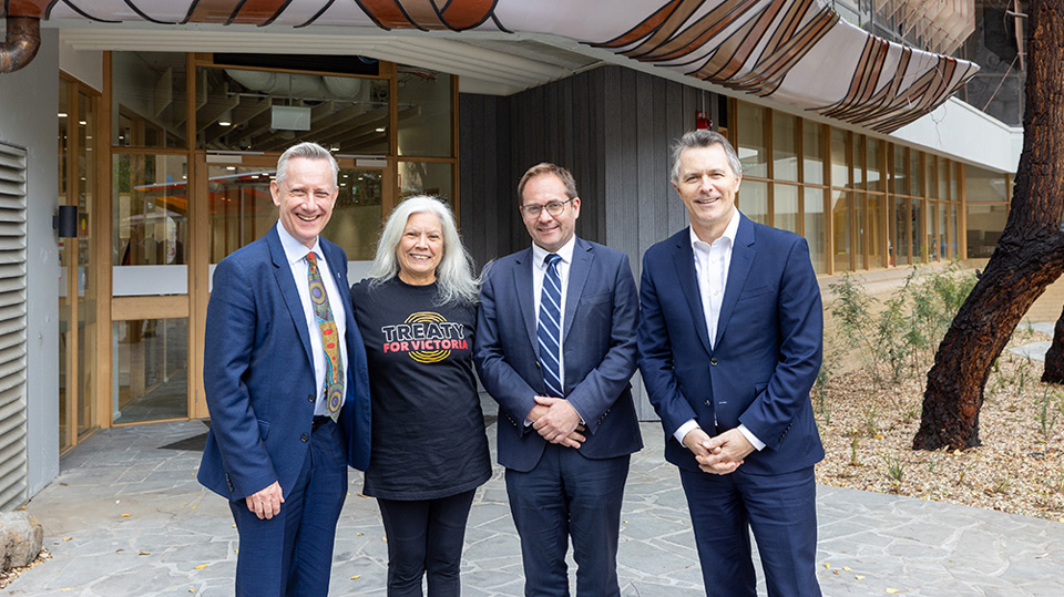 Three men in suits stand with a woman wearing a Treaty t-shirt in front of an Indigenous design over a doorway