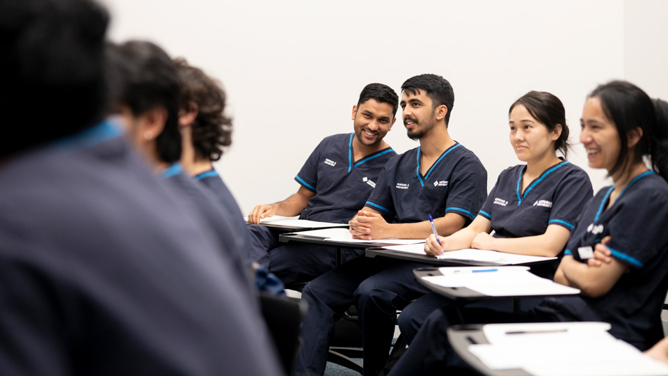 Students sitting in a room looking at the front. One student is leaning towards another student and smiling.