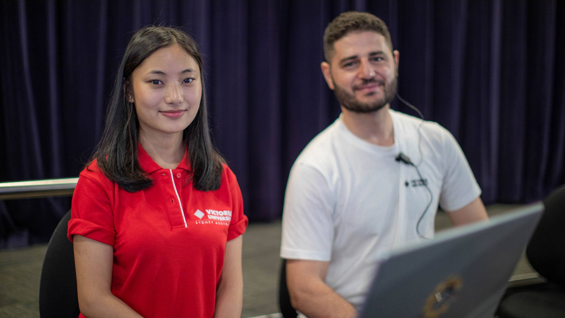 A VU Sydney student in a red shirt uniform, sitting next to a student. Both are looking at the camera and smiling.