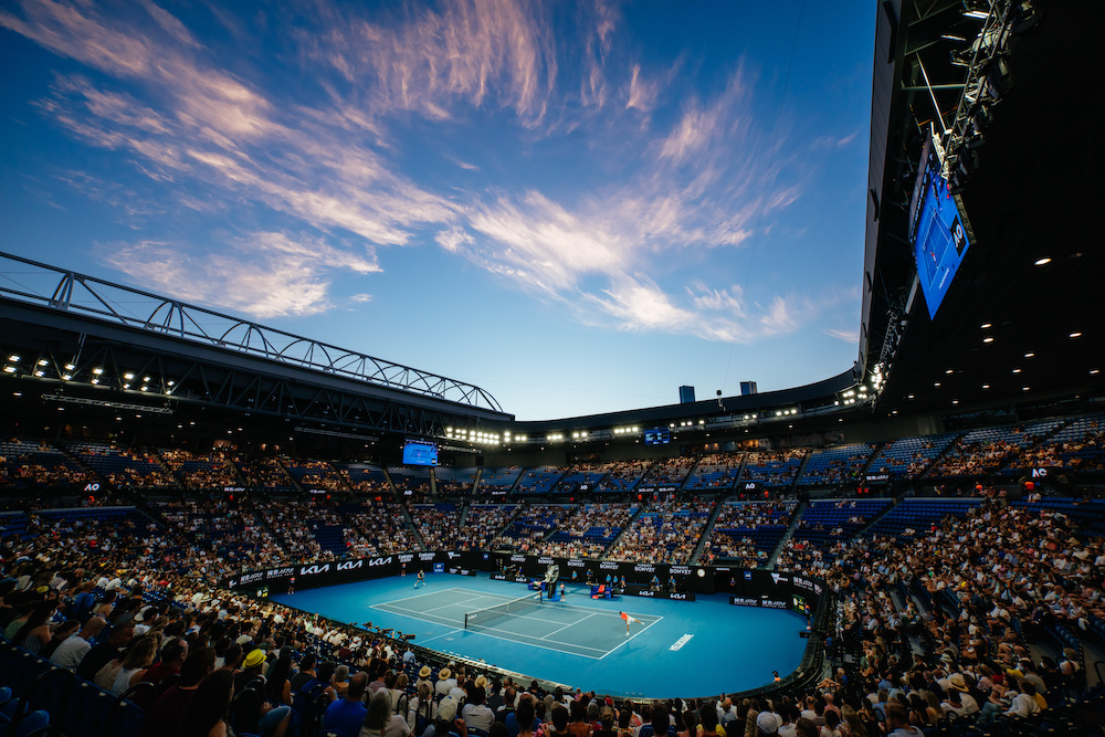 A tennis court arena. The arena is filled by a crowd of spectators. The sky is dark blue, as the sun is setting. The court is illuminated by artificial lights.