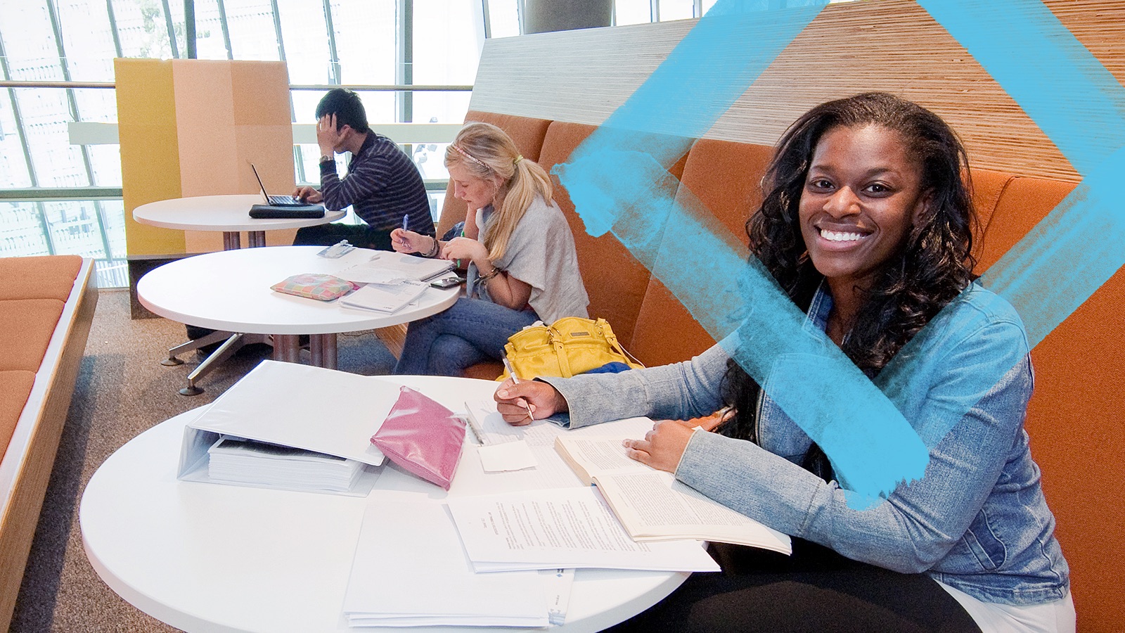A smiling student studying in the library's study area, with two other students in the background.