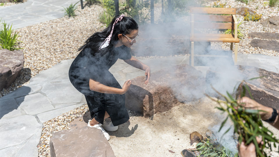 An attendee at a Smoking Ceremony at the Moondani Balluk garden. The person is crouching down close to the fire pit.