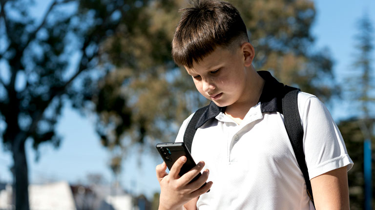 Teenage boy standing outside, holding and looking at his phone, with a concerned expression on his face.