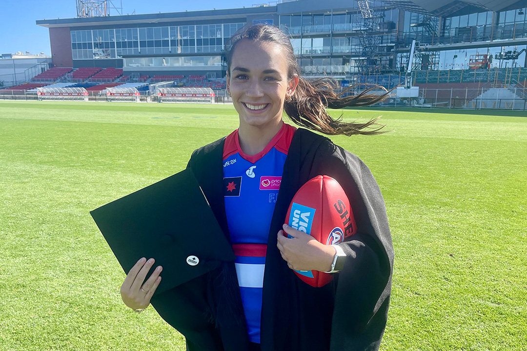 A young woman in a bule, white and red sport uniform and graduation gown, holds a football