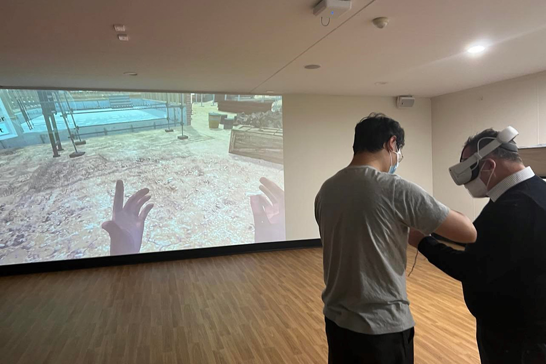 A man in a VR handset collaborates with another man in front of a large VR screen with a building site