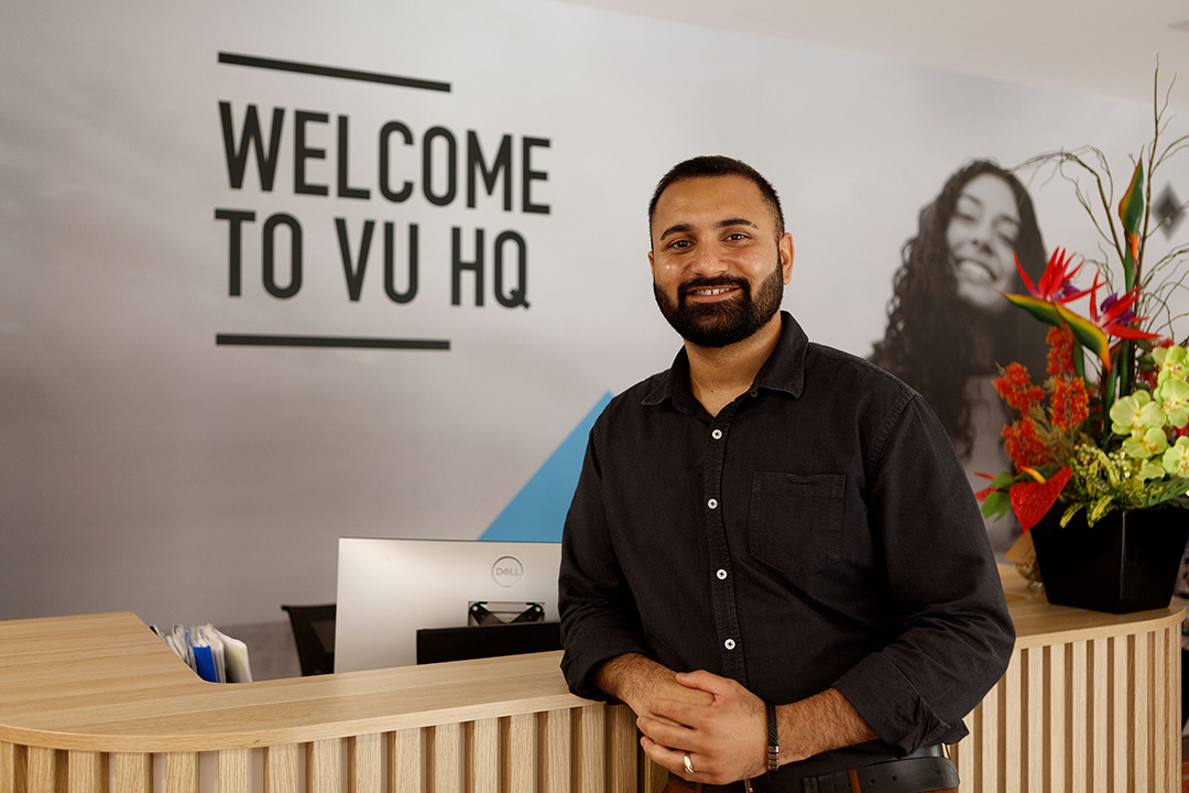 A smiling man leans on a reception desk. On the wall behind is written 'Welcome to VU HQ'