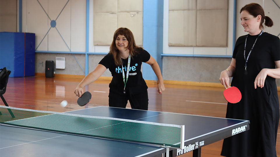 Two people play table tennis. One is wearing a thrive* shirt.
