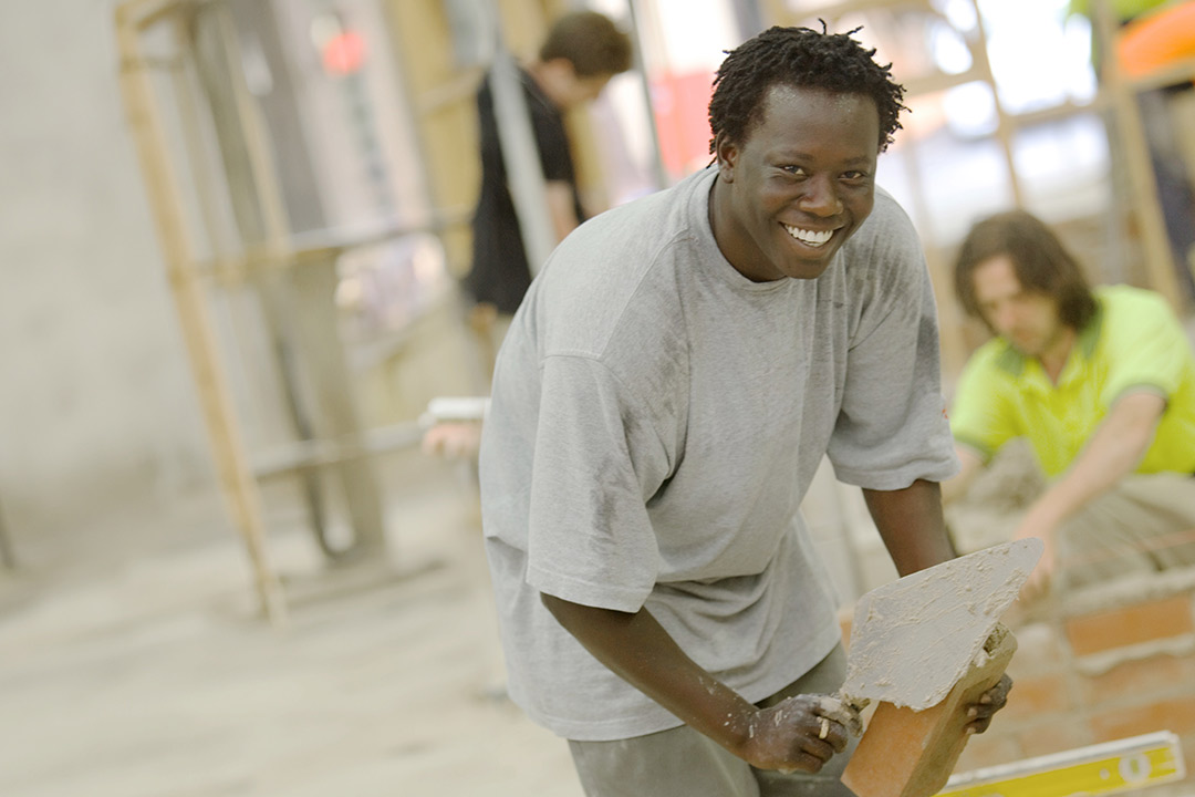 A young man holds a trowl and smiles while laying bricks in a large workspace