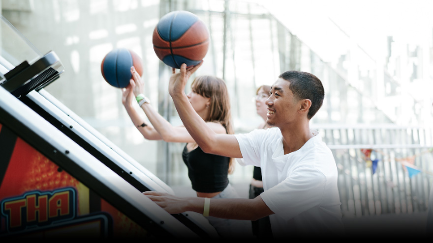 Two students about to throw a basketball into a basketball arcade machine. One student is watching in the background.