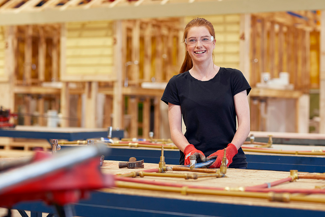 A smiling young woman wears safety goggles and gloves while working in a large carpentry workshop