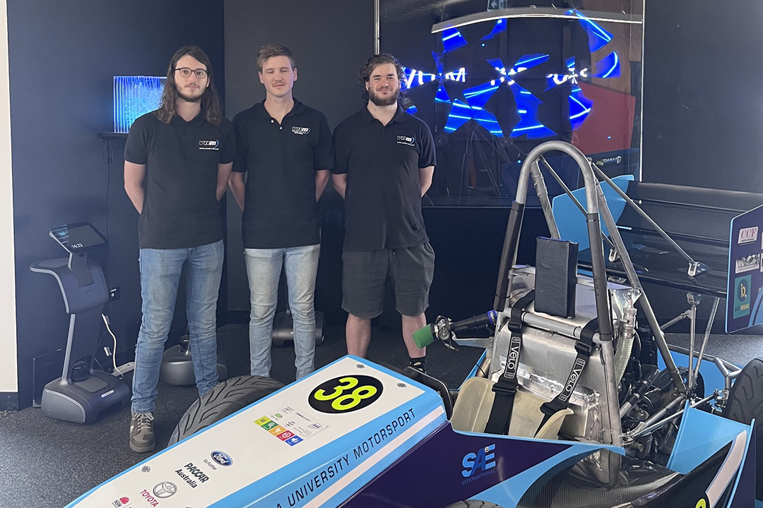 3 men in VU t-shirts pose in a room with a digital screen and a VU branded race car