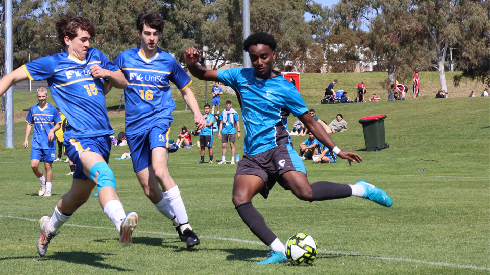 A VU Vultures football team member kicks the ball while being chased by two opponents - UniSport Nationals 2024