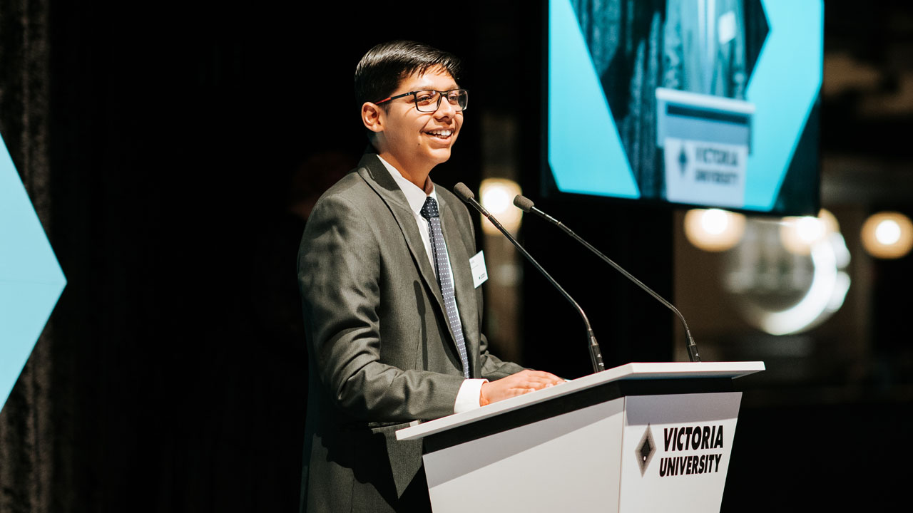 A student in formal wear standing on stage behind a podium. Podium has Victoria University written on it.