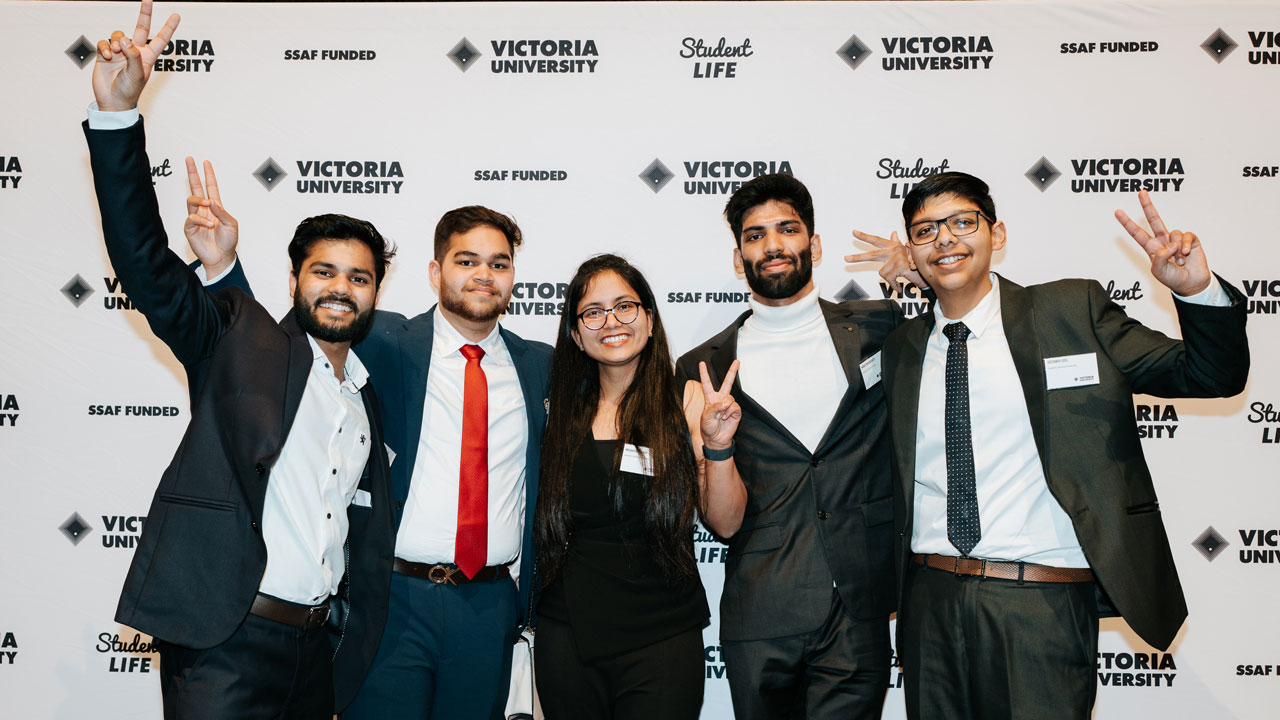 Students in formal wear standing in front of a VU backdrop. Students are holding up peace signs with their fingers.