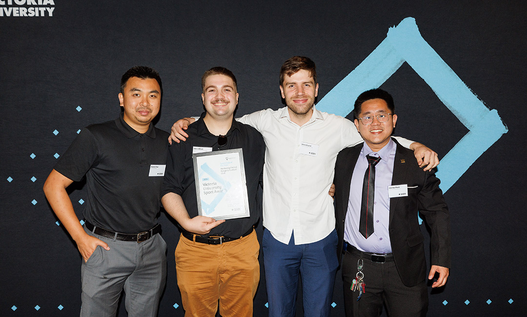 Four young men in semi-formal dress pose with an award in front of a Victoria University banner