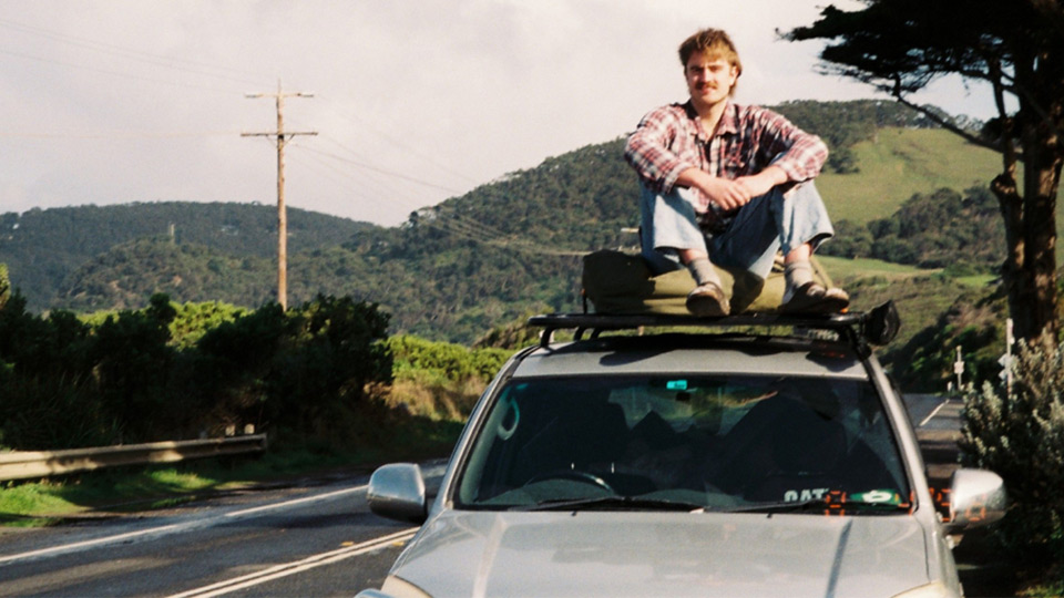 Duncan James smiles at the camera, sitting on the roof of a parked car alongside the Great Ocean Road. 