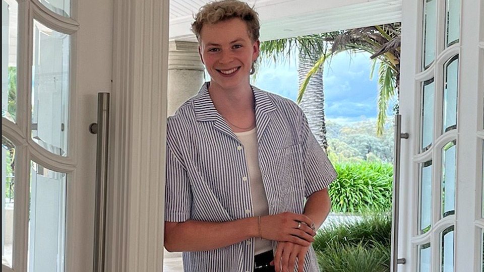 Lachy Moroney smiles at the camera. He is standing in front of an open door in a tropical landscape.