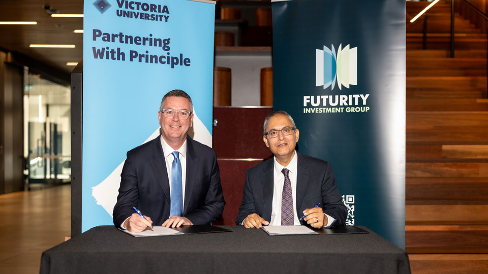 Two men sitting behind a desk. Both have finished signing a document in front of them. Behind them are banners with the Victoria University and Futurity logos.