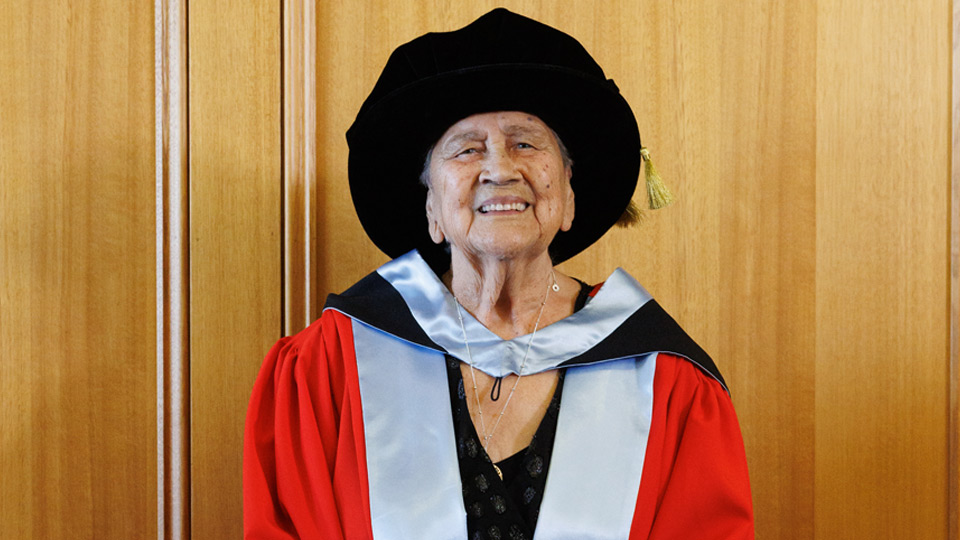 Dr Alma Thorpe smiling, standing in front of a wood panelled wall, - Alma is wearing her Honorary Doctorate gown and cap