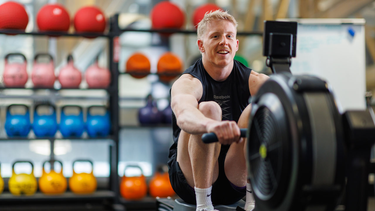 A person in gymwear working out on a rowing machine. Behind them is a shelf full of kettleballs.