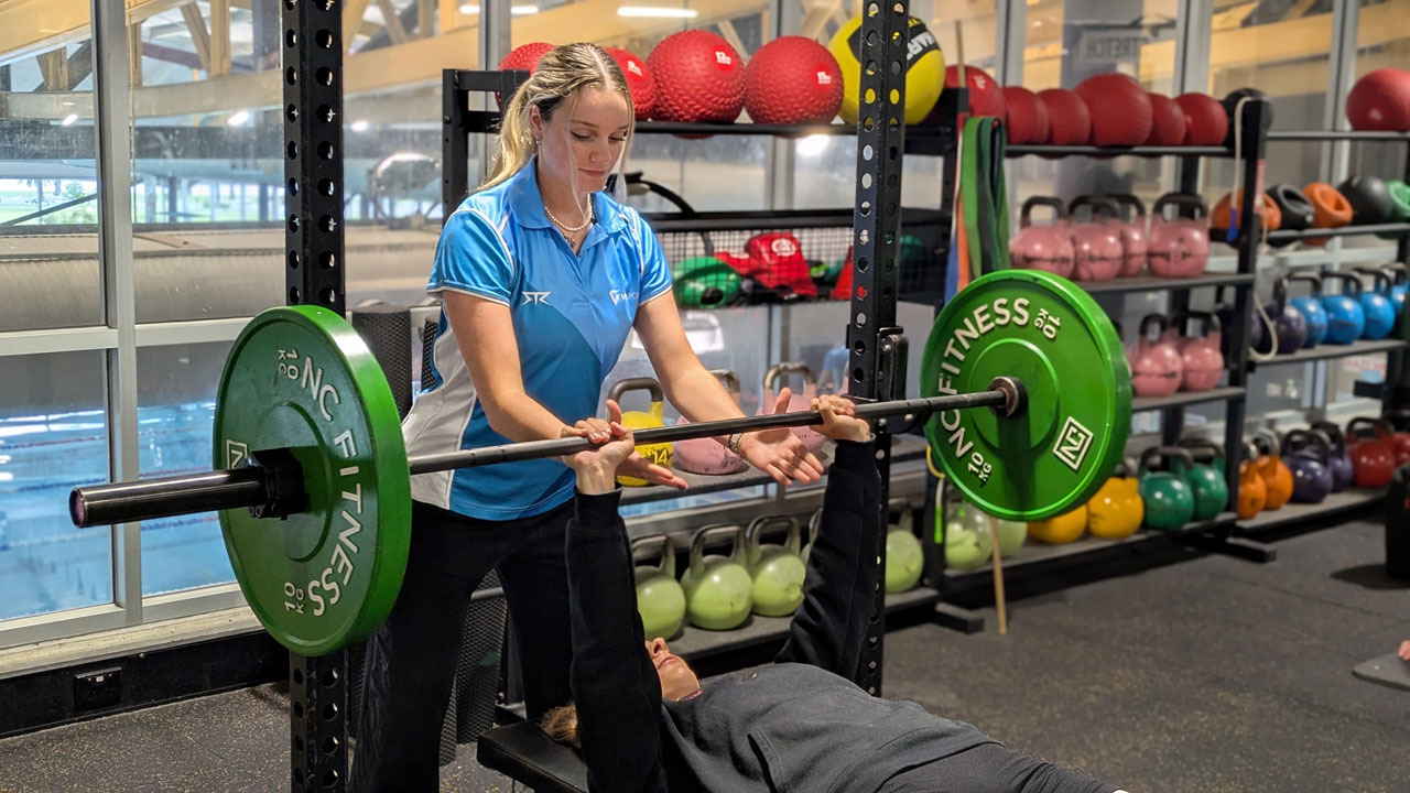 A person benching at a gym. Behind them is a personal trainer holding the bar for support.