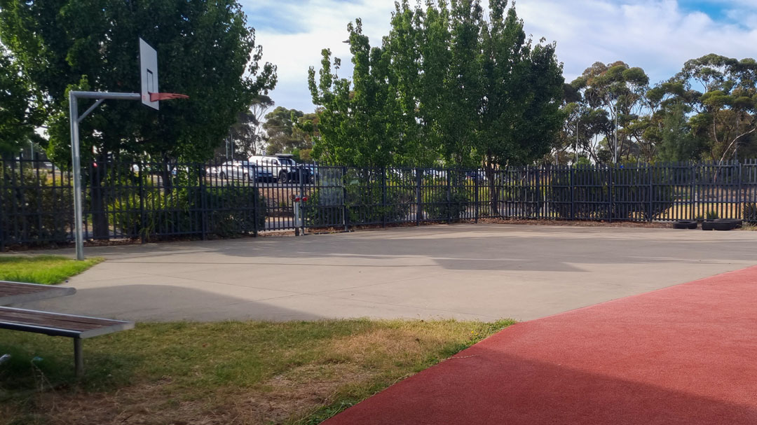 An outdoor concrete court with a basketball hoop at one end. Next to it is a red field for training and exercise.