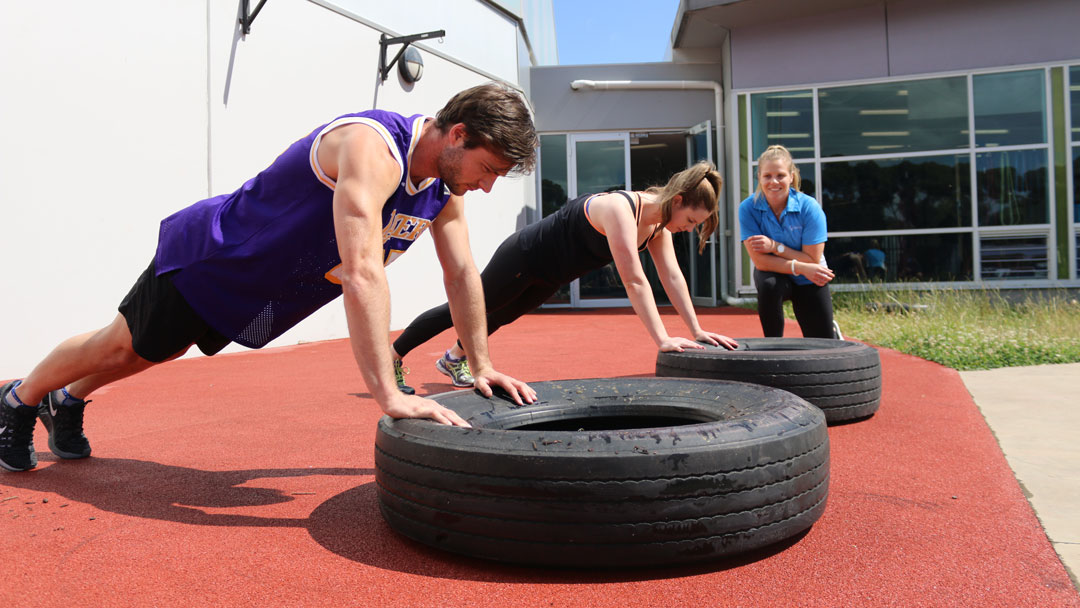 Two people outside in a push-up postion, with their hands on top of a tyre. A personal trainer is leaning and observing them in the background.