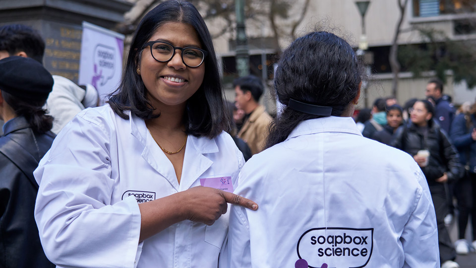 Dr Fernando smiles at the camera at a Soapbox Science event, pointing out the event logo on the back of a fellow attendee