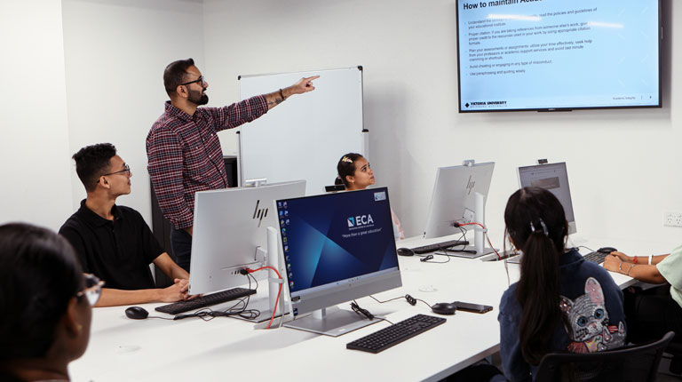 Students sitting behind computers. A teacher is standing up and pointing at a presentation on the big screen.
