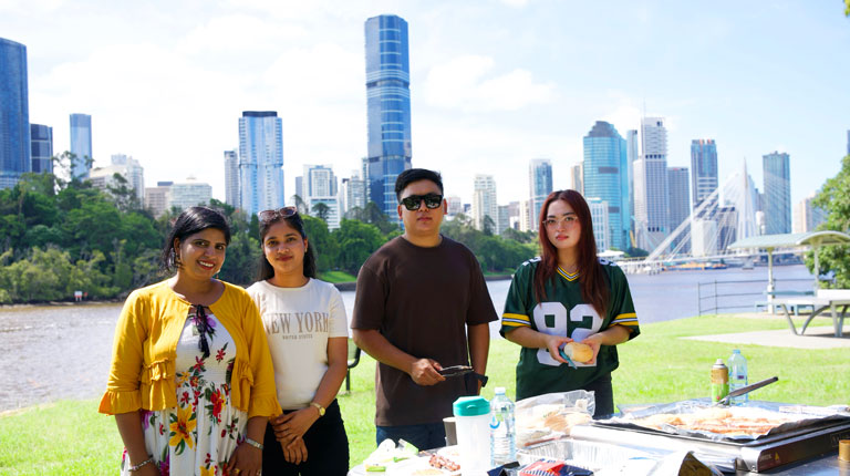 Four students standing outside in front of a barbeque. Behind them is the Brisbane river and city buildings.