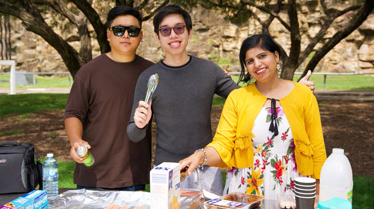 Three VU students standing behind an outdoor public barbeque. Behind them is a park with tall trees.