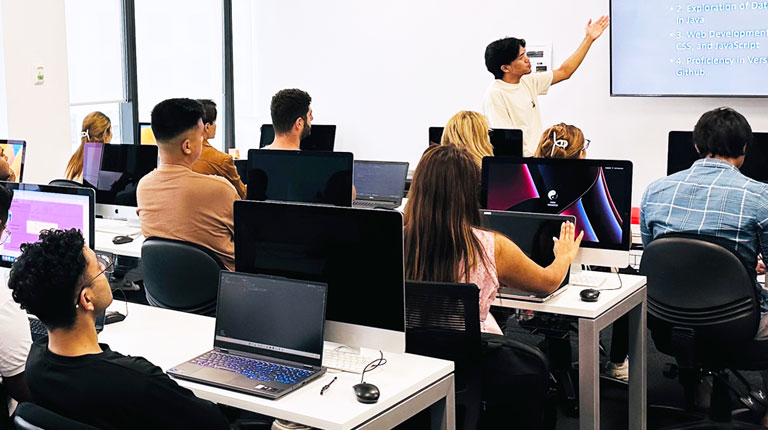 A group of students sitting at computers. One student is at the front of the room pointing to code on a projector.