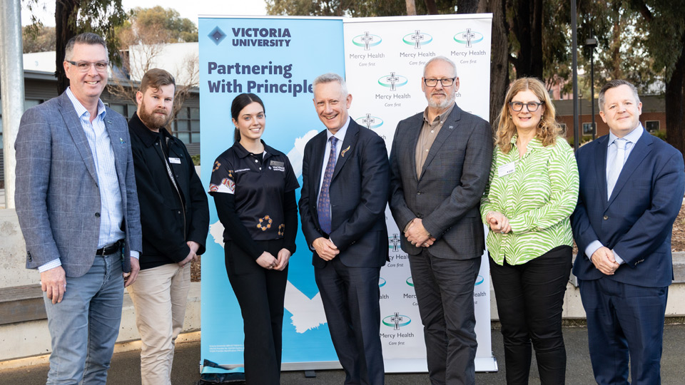 L-R: The Hon. Wade Noonan, David Schreurs, Bridget McKenna, Professor Adam Shoemaker, Wayne Butson, Professor Gina Kruger, Professor Andy Hill stand outside, smiling in front of VU and Mercy Health pull-up banners