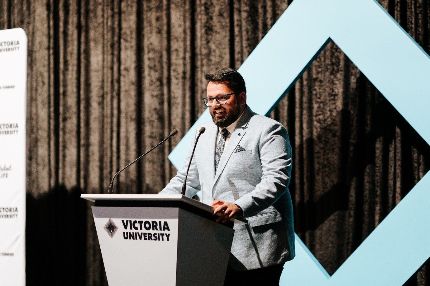 A man in a crisp grey suit speaks at a Victoria University branded podium, in front of theatre curtains
