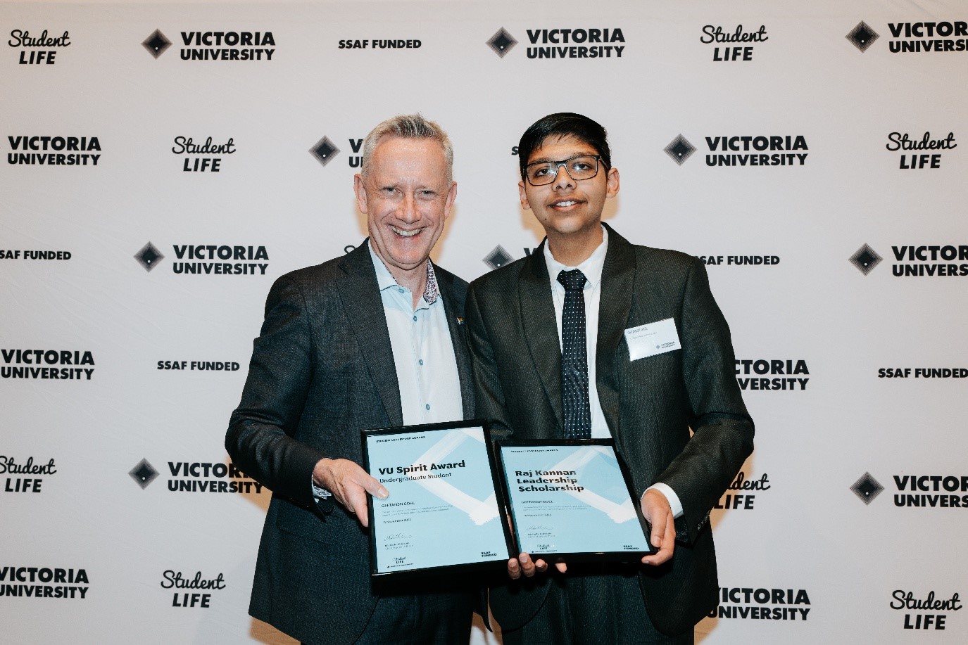 A middle-aged man in a suit holds an award and smiles with a young man in a suit, in front of Victoria University logos