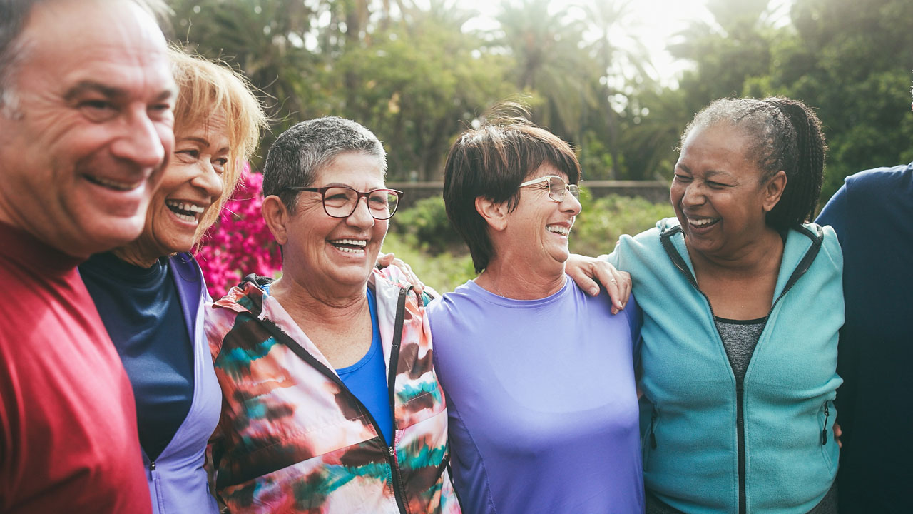 A group of senior people standing outside in a park, arms over each others' shoulders.
