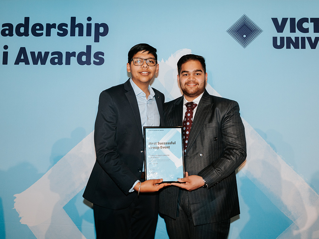 Two young men in smart suits pose with an award in front of a Victoria University banner