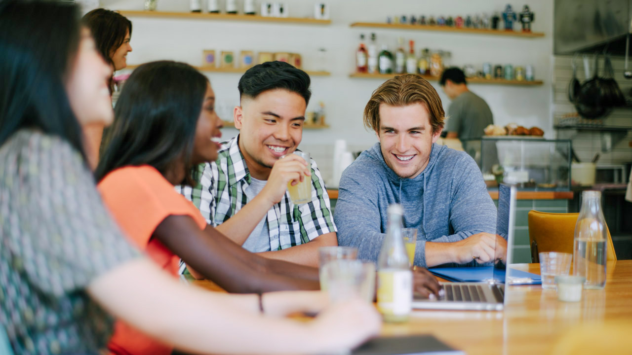 Four students sitting inside a cafe sharing a laptop smiling and laughing together.