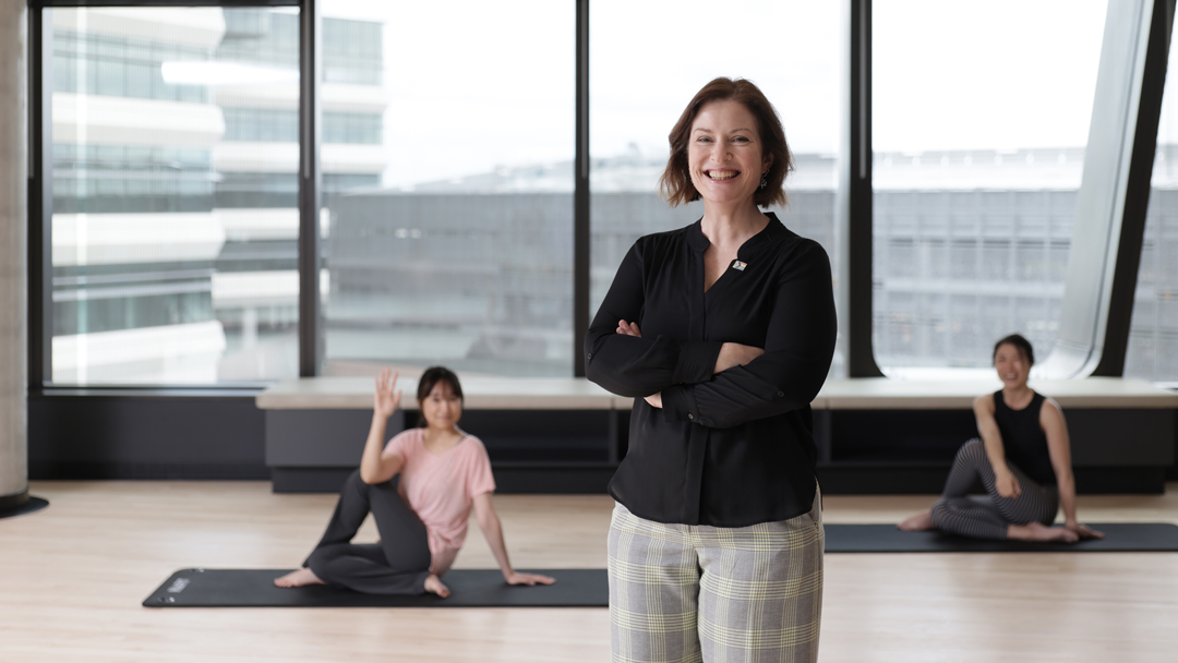 A woman in professional dress poses in youga class held in a large room with tall glass windows