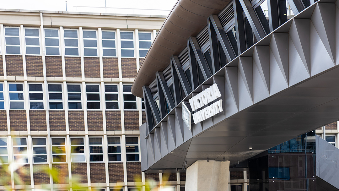 A modern over-road bridge with 'Victoria University' logo leads to a 1970s campus