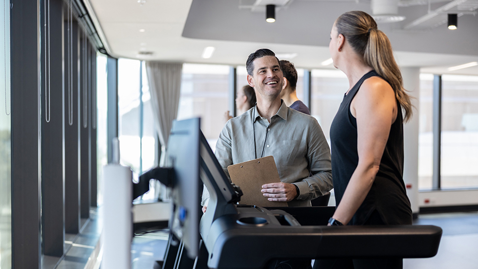 A man holding a clipboard speaks to a woman on a treadmill in a modern gym with large windows