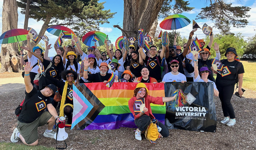 A group of smiling people in an Australian park with rainbow umbrellas, pride flags and Victoria University banners
