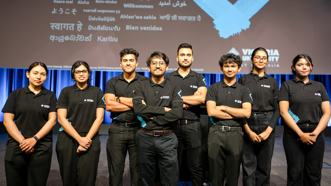 A multicultural group of friendly young adults in black staff uniform in front of a welcome banner with different languages and Victoria University branding