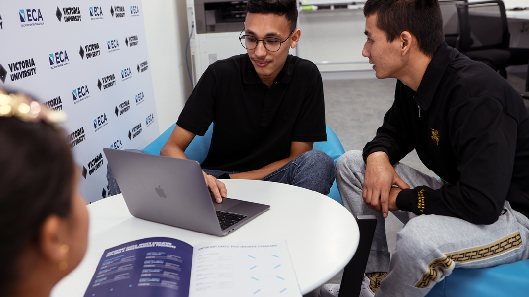 Two students seated at table, in conversation looking at laptop