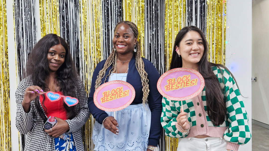 Three smiling students at fun event, holding colorful 'Block besties' signs, standing in front of decorated wall. 