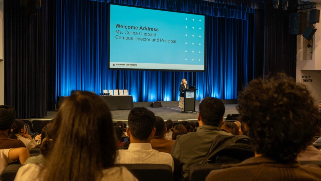 A speaker stands on stage presenting a blue PowerPoint slide to a large audience seated in a theatre at VU Sydney.