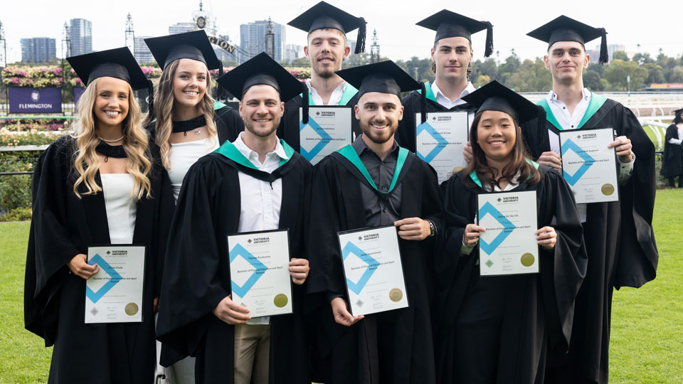 A group of 8 graduates smile proudly after their graduation ceremony at Flemington Racecourse